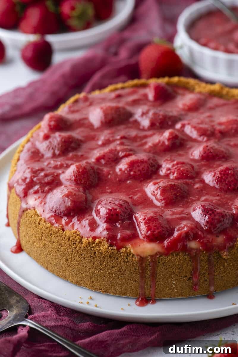 Strawberry Cheesecake Recipe with Fresh Strawberry Topping 20 A cheesecake with a graham cracker crust, topped with a glossy strawberry sauce and whole strawberries, sits on a white plate.