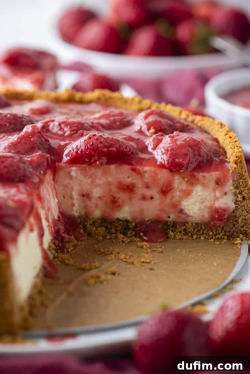 Strawberry Cheesecake Recipe with Fresh Strawberry Topping 5 A close-up of a strawberry cheesecake with a graham cracker crust. A slice has been removed, showing creamy filling and strawberry topping. Fresh strawberries are scattered around the cheesecake.