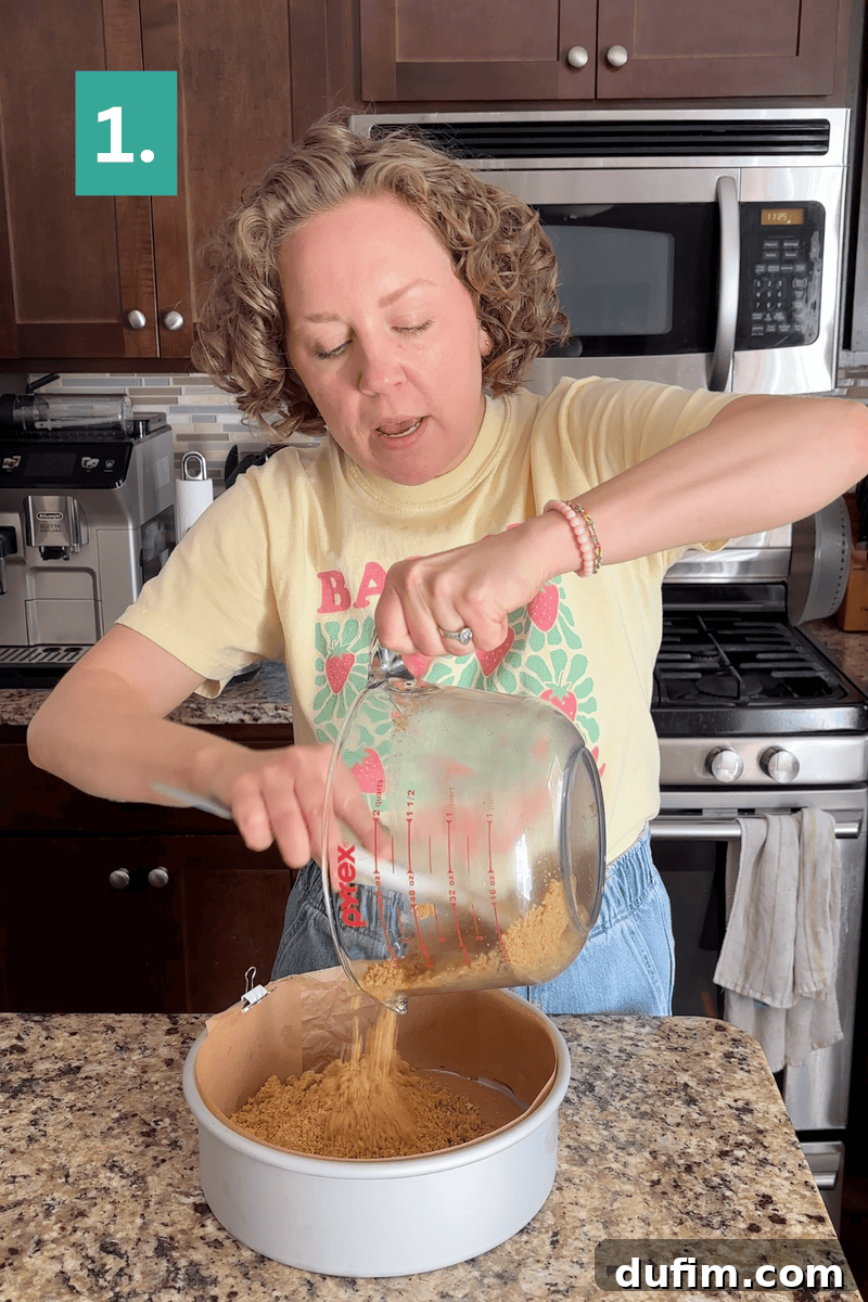 Strawberry Cheesecake Recipe with Fresh Strawberry Topping 6 A woman pours crushed graham crackers from a glass measuring cup into a round baking pan on a kitchen counter.
