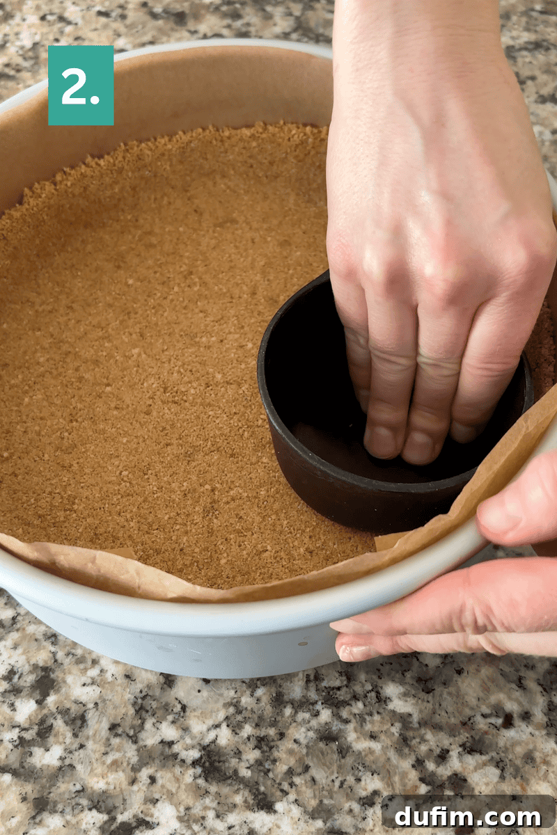 Strawberry Cheesecake Recipe with Fresh Strawberry Topping 7 A person presses a measuring cup into a pie pan lined with parchment and filled with a crumb crust, shaping it evenly.