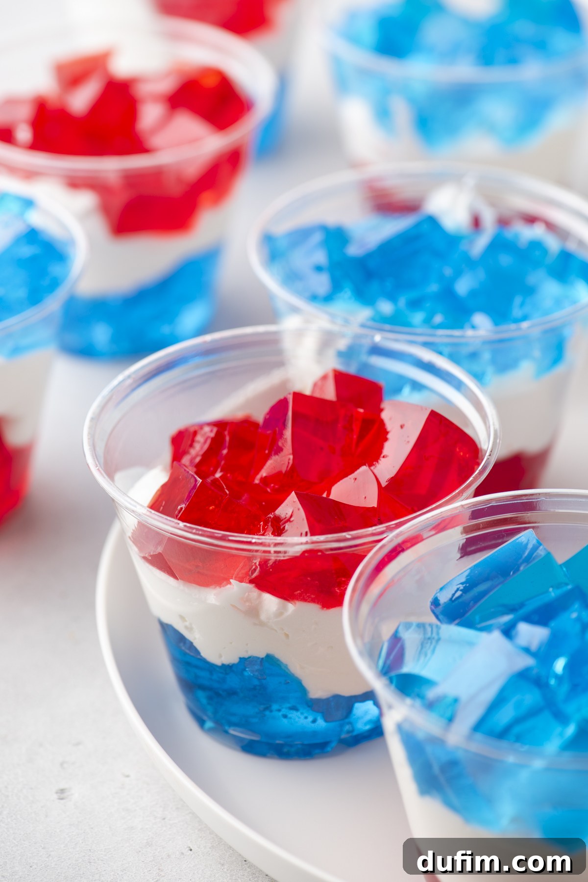 Clear plastic cups filled with layered red, white, and blue gelatin are arranged on a white plate, with the vibrant colors creating a festive and patriotic dessert display.