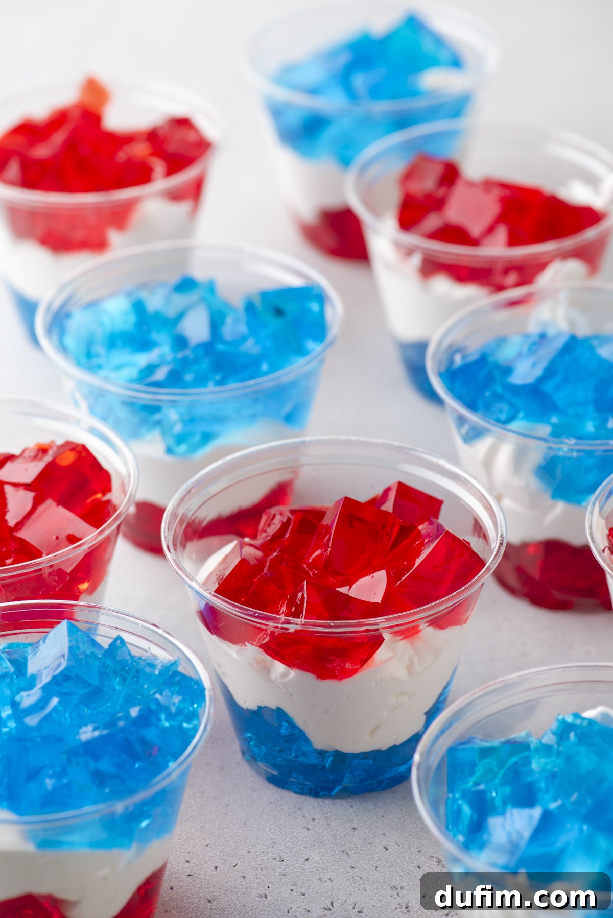 Plastic cups filled with layered red and blue gelatin and white cream, creating a patriotic, tricolor dessert. The cups are arranged on a light surface.