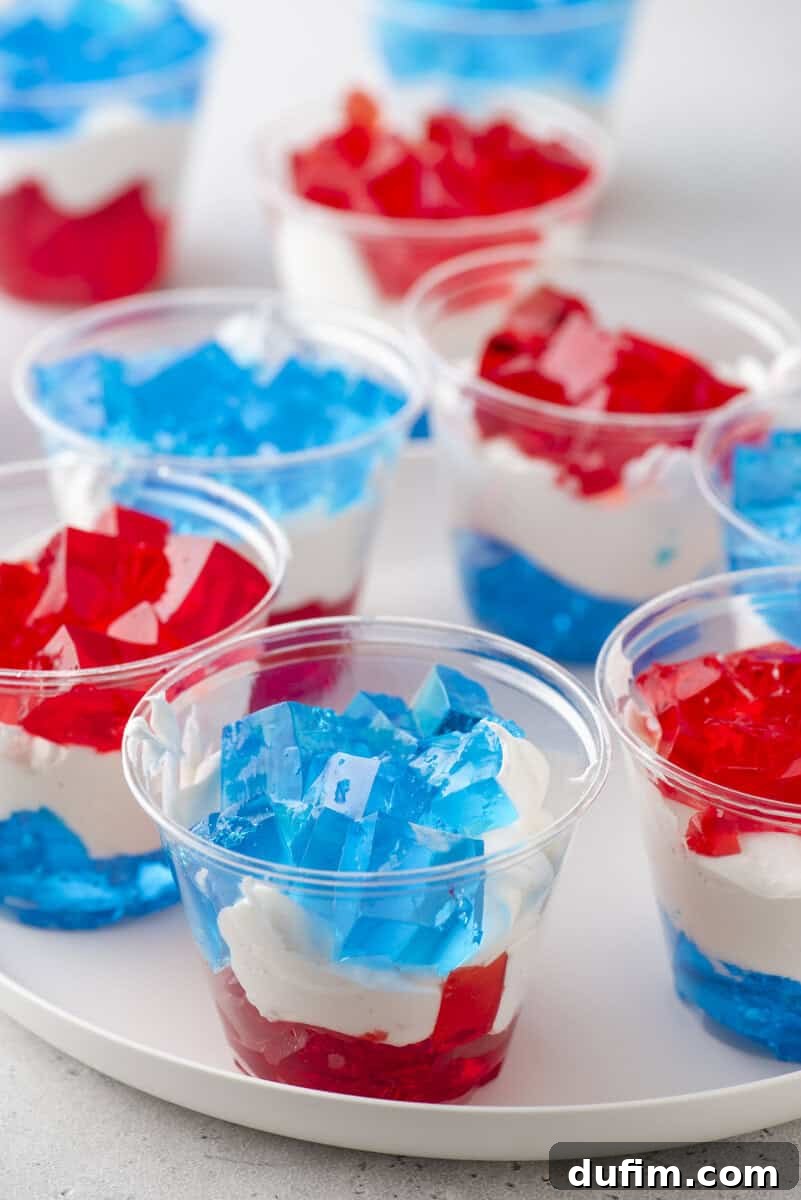 Clear plastic cups filled with layered red and blue gelatin and white whipped cream, arranged on a white tray. The layers create a festive, colorful dessert, suggesting a patriotic or celebratory theme.