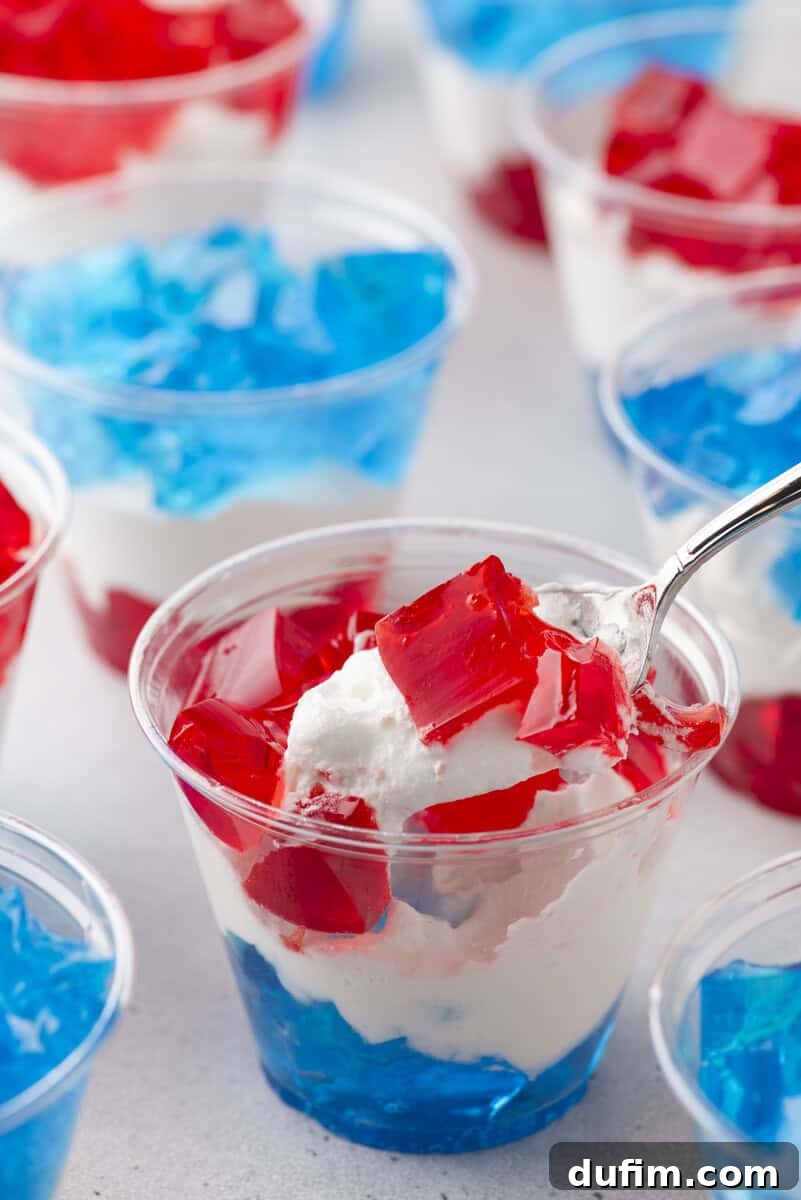 Plastic cups filled with layered red and blue gelatin and white whipped cream. A spoon is lifting a portion from one cup, showing the colorful dessert layers. Other cups are visible in the background.
