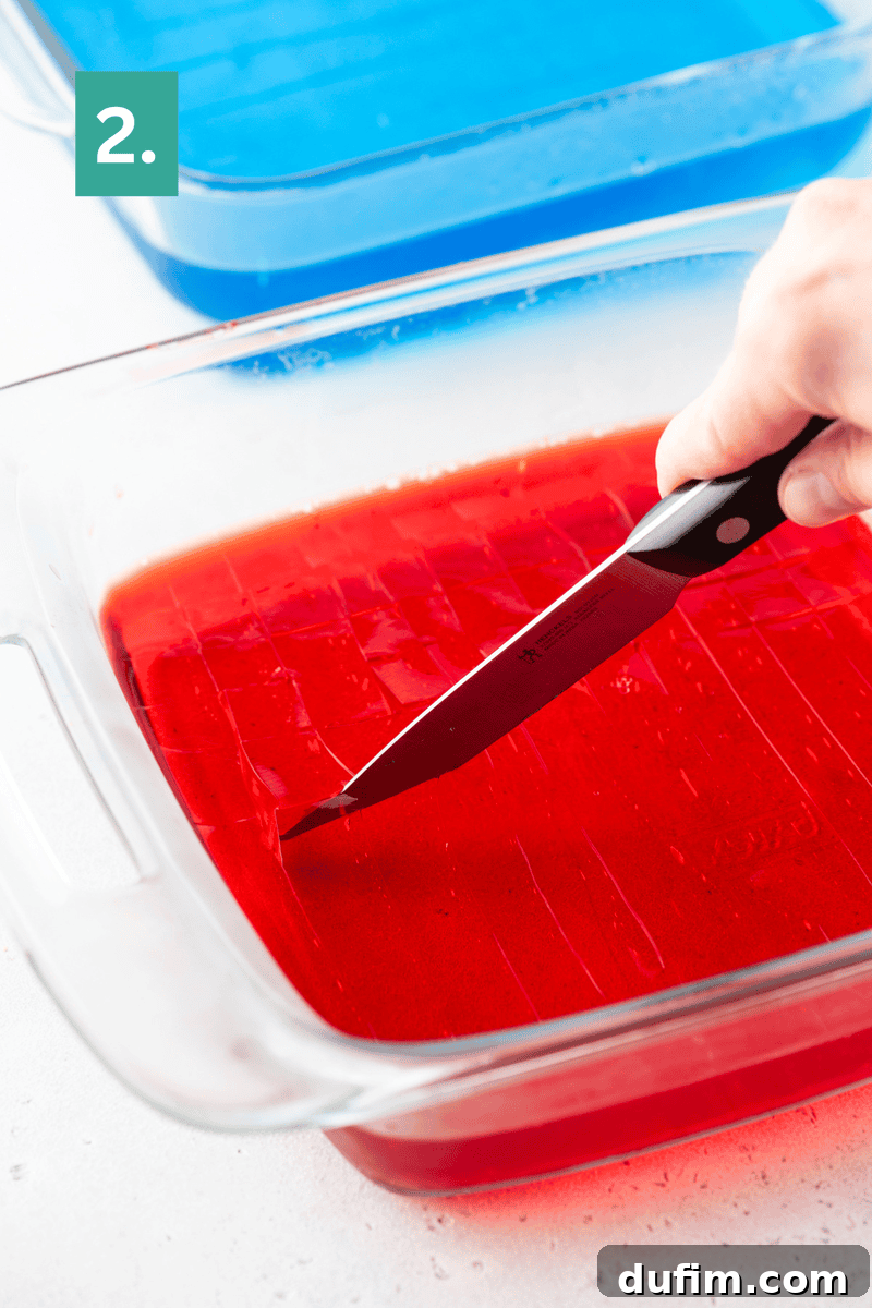 A hand cuts red gelatin in a glass dish with a knife; a dish of blue gelatin sits in the background.