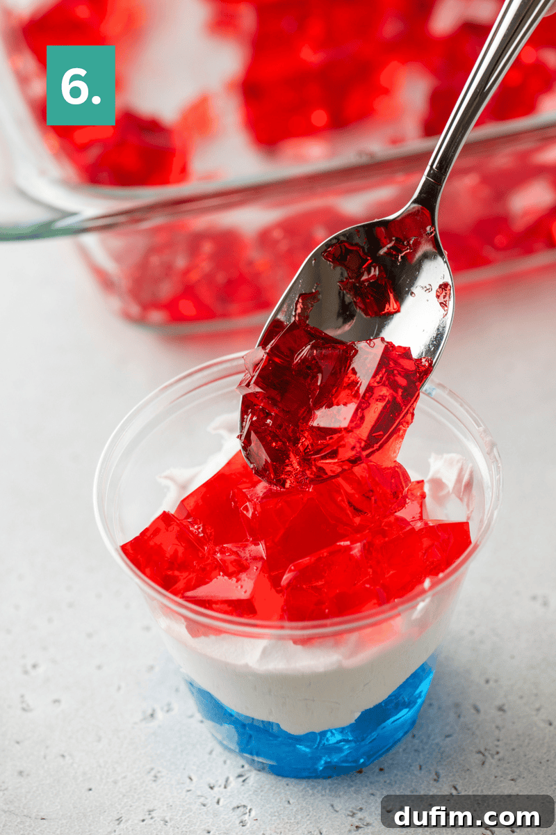 A spoon adds red gelatin cubes on top of whipped cream and blue gelatin in a clear cup.