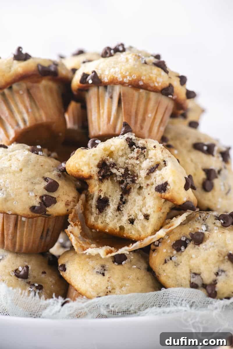 A close-up of a plate stacked with chocolate chip muffins, with one muffin in the center split open to show the soft, chocolate-filled interior. The muffins are sprinkled with sugar crystals.