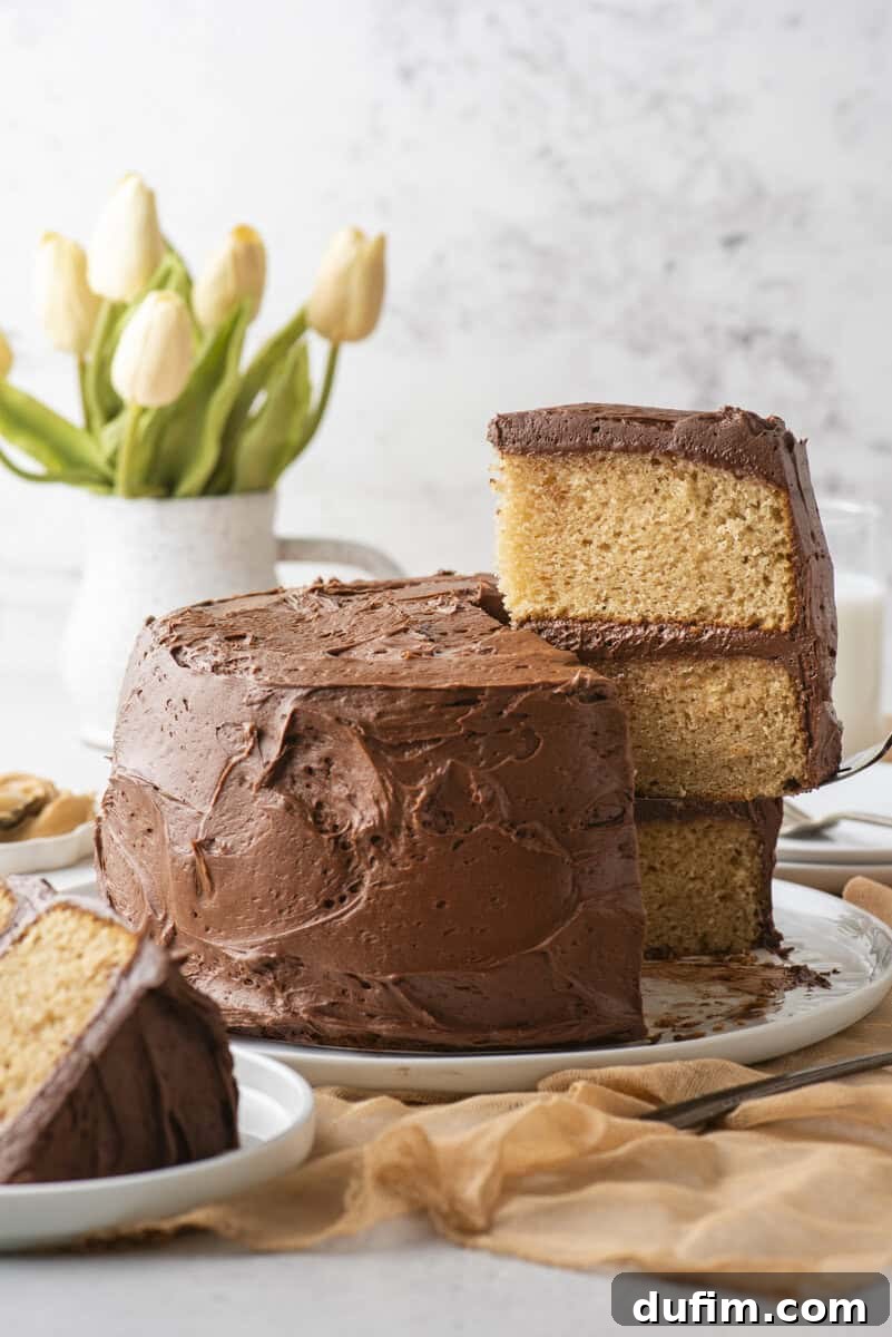 Peanut Butter Cake with Chocolate Frosting Recipe 2 A two-layer yellow cake with chocolate frosting sits on a plate, with one slice lifted up. A vase of yellow tulips and a glass of milk are in the background.