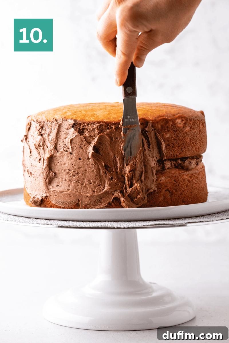 Peanut Butter Cake with Chocolate Frosting Recipe 14 A hand uses a spatula to spread chocolate frosting on the side of a two-layer yellow cake, placed on a white cake stand.