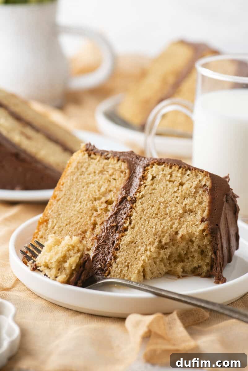 Peanut Butter Cake with Chocolate Frosting Recipe 16 A slice of yellow cake with chocolate frosting sits on a white plate with a fork. A glass of milk and more cake slices are in the background.
