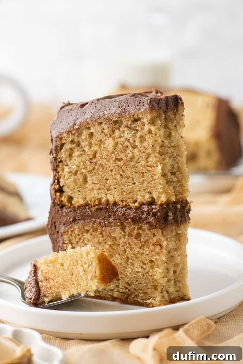 Peanut Butter Cake with Chocolate Frosting Recipe 3 A slice of two-layer yellow cake with chocolate frosting sits on a white plate. A fork holds a bite-sized piece in front of the cake. The background is softly blurred.