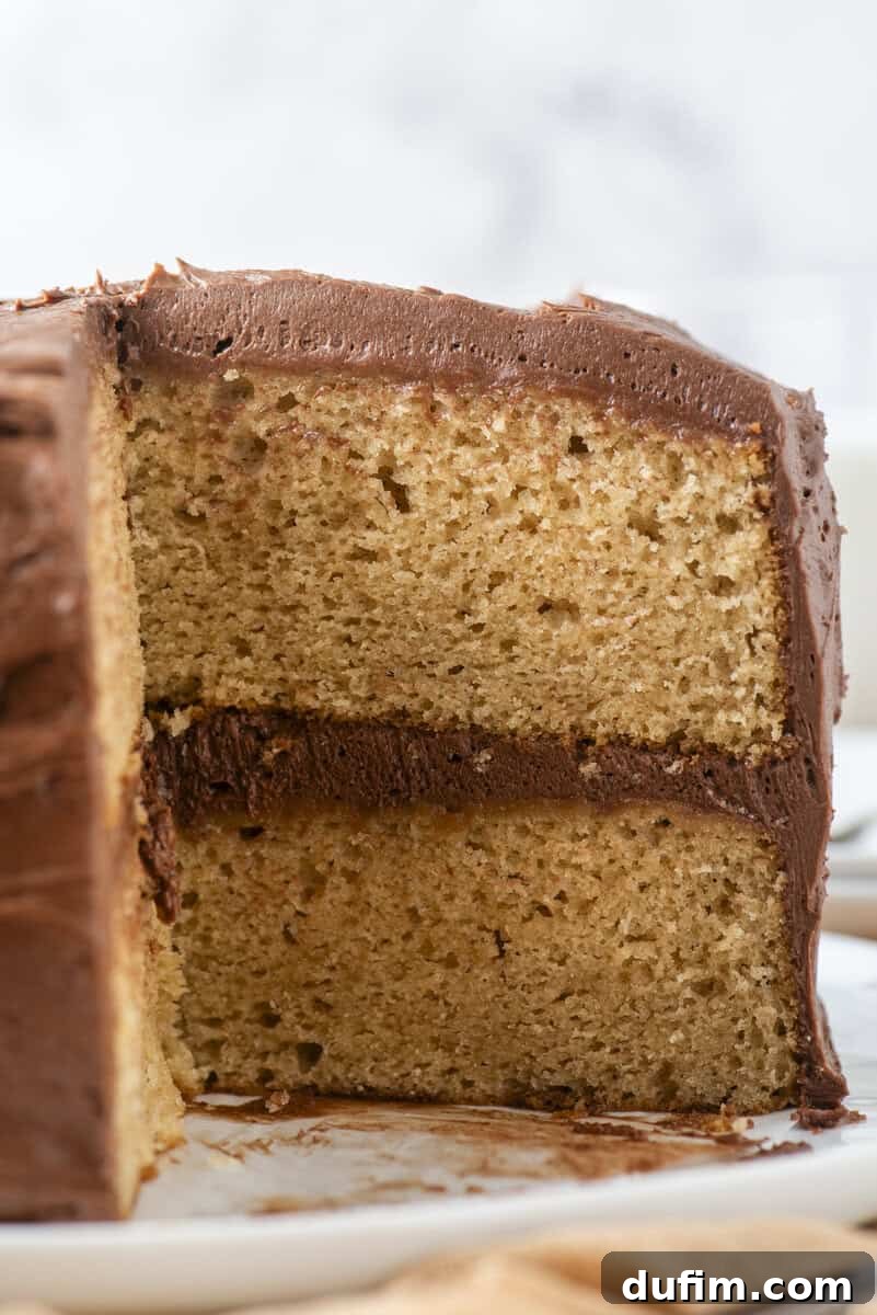 Peanut Butter Cake with Chocolate Frosting Recipe 5 A close-up of a sliced yellow cake with two layers, filled and topped with chocolate frosting, on a white plate.