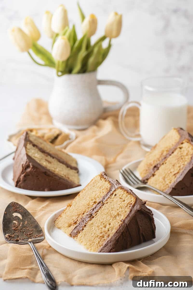 Peanut Butter Cake with Chocolate Frosting Recipe 6 Three slices of yellow cake with chocolate frosting on white plates, forks beside them, with a white mug of milk and a vase of yellow tulips in the background on a peach-colored cloth.