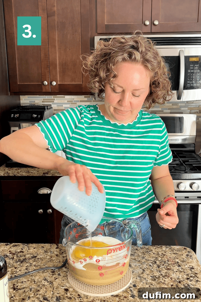 Peanut Butter Cake with Chocolate Frosting Recipe 9 A woman in a green striped shirt pours liquid from a measuring cup into a Pyrex bowl with eggs and sugar on a kitchen counter.