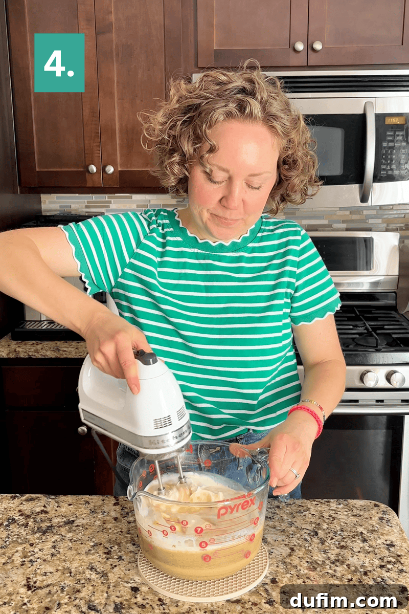 Peanut Butter Cake with Chocolate Frosting Recipe 10 A woman in a green striped shirt uses a hand mixer to blend ingredients in a Pyrex bowl on a kitchen counter.