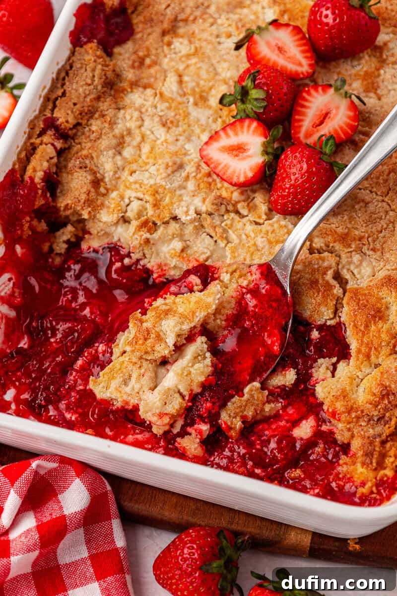 A close-up of a strawberry cobbler in a white baking dish, with a spoon serving some. Fresh strawberries and a red checkered cloth are nearby.