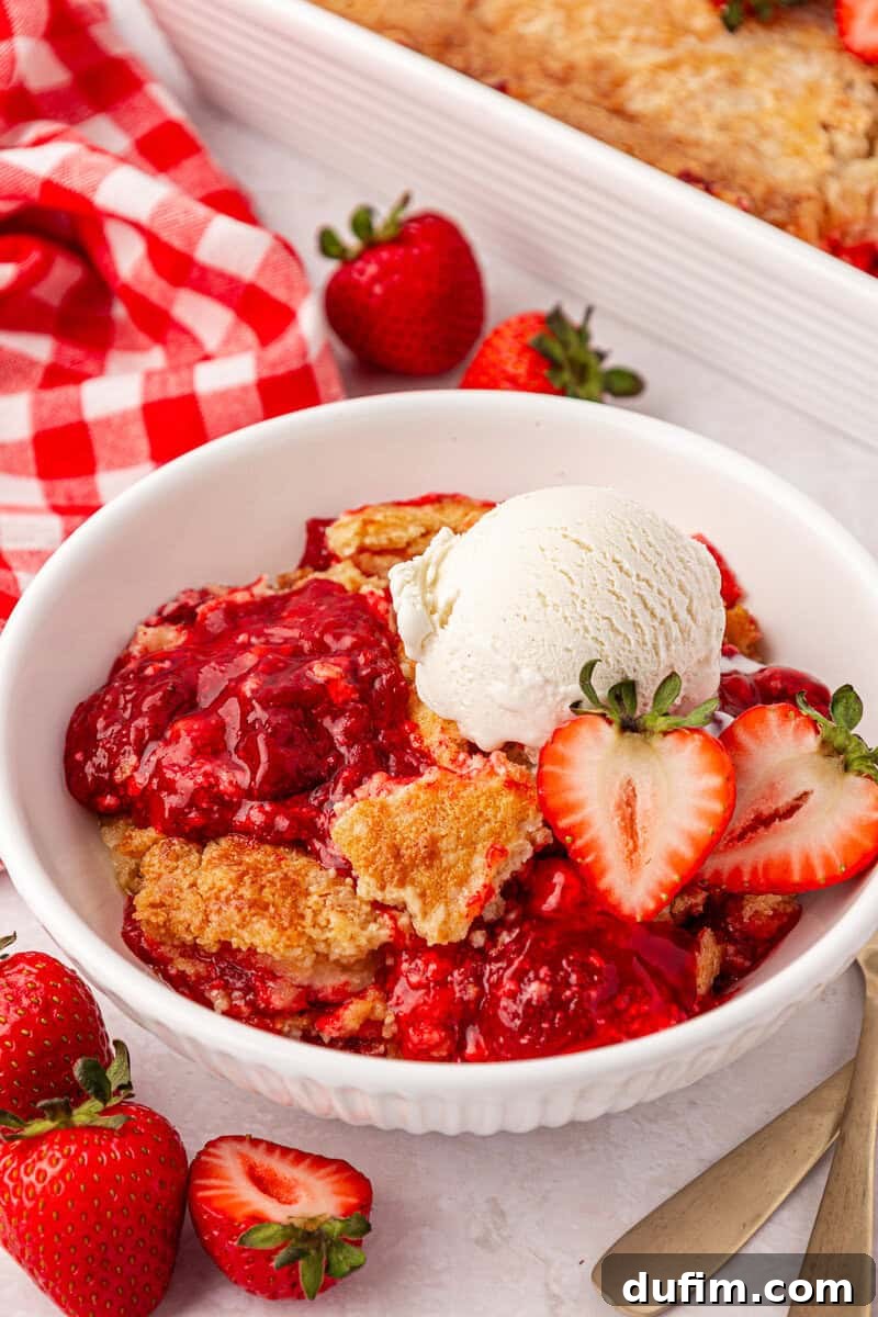 A bowl of strawberry cobbler topped with a scoop of vanilla ice cream and sliced strawberries, with fresh strawberries and a red checkered napkin nearby.