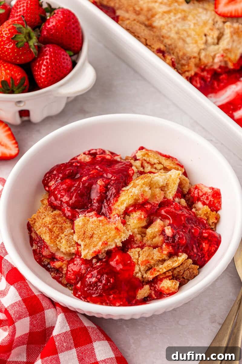 A white bowl filled with strawberry cobbler sits next to a dish of fresh strawberries and a red checkered napkin on a white surface.