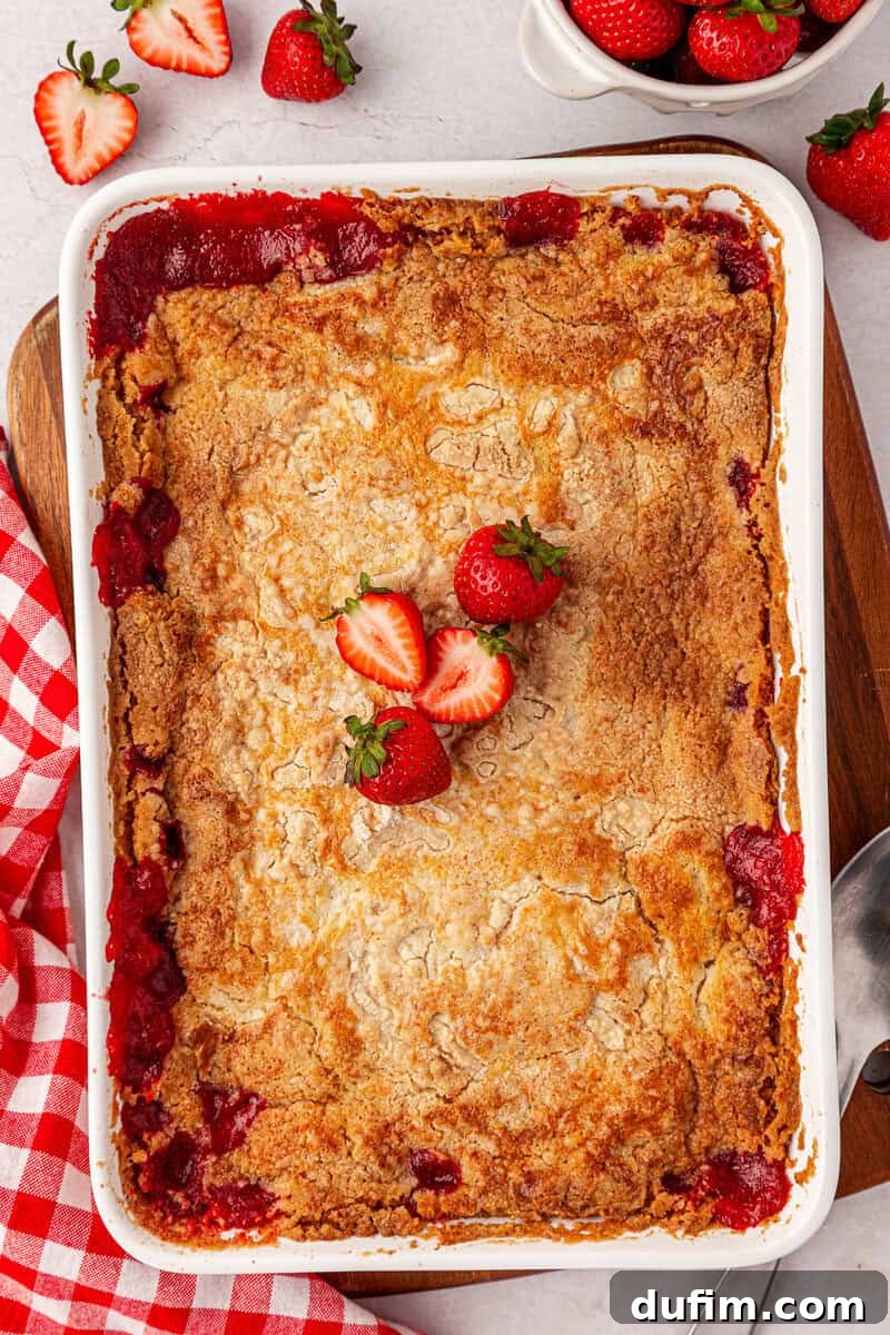 A baked strawberry cobbler in a white dish, topped with three fresh strawberries, sits next to a red checkered cloth and strawberries on a white surface.
