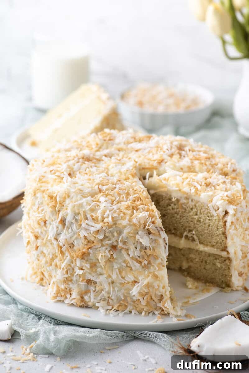 Homemade Coconut Cake That Tastes Like Real Coconut 2 A round layer cake covered in toasted coconut flakes sits on a white plate, with one slice cut and slightly pulled out. In the background are a glass of milk, a bowl of coconut, and a vase of flowers.
