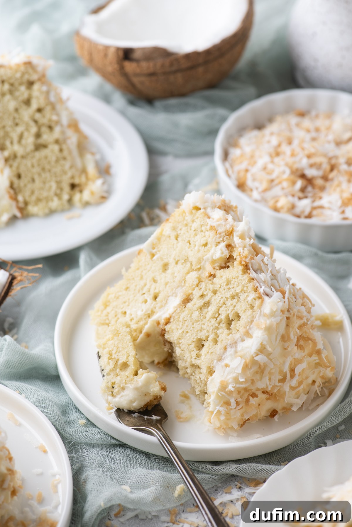 Homemade Coconut Cake That Tastes Like Real Coconut 10 A slice of coconut cake with creamy frosting and toasted coconut flakes sits on a white plate, with a fork beside it.