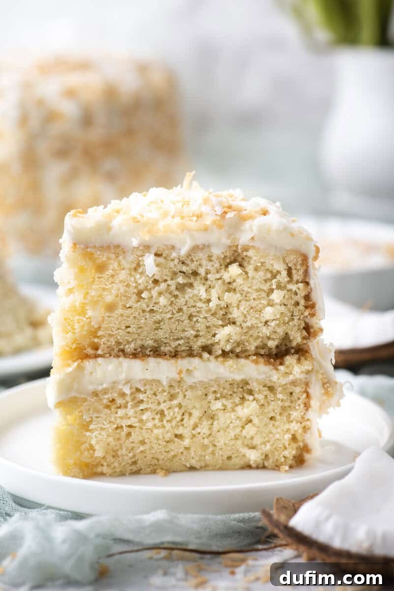 Homemade Coconut Cake That Tastes Like Real Coconut 11 A close-up of two stacked slices of coconut cake with white frosting and toasted coconut flakes on top, served on a white plate.