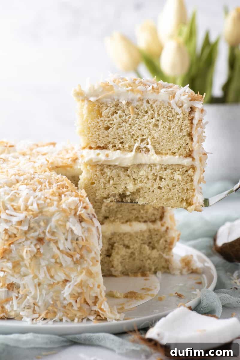 Homemade Coconut Cake That Tastes Like Real Coconut 3 A slice of coconut layer cake with cream filling and toasted coconut flakes is being lifted from the whole cake. White tulips and a coconut shell are in the background.