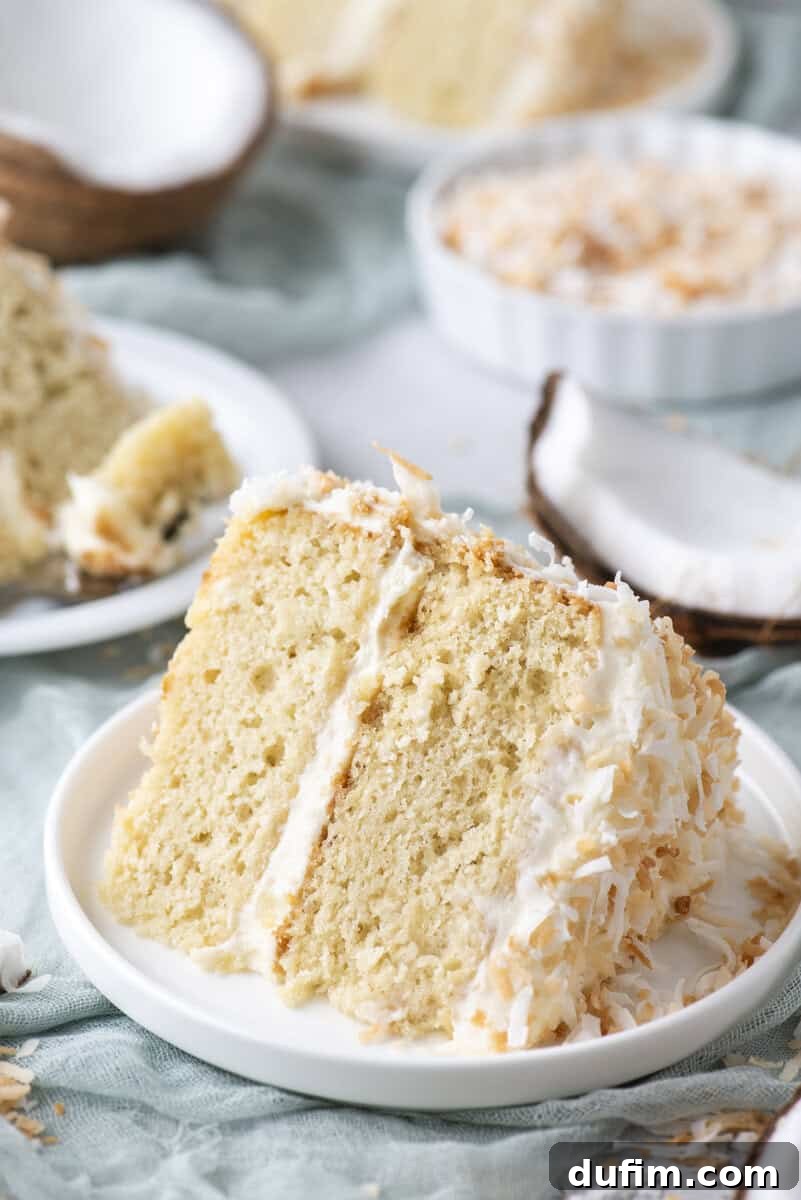 Homemade Coconut Cake That Tastes Like Real Coconut 12 A slice of coconut layer cake with white frosting and shredded coconut sits on a white plate, with more cake and coconut pieces in the background.