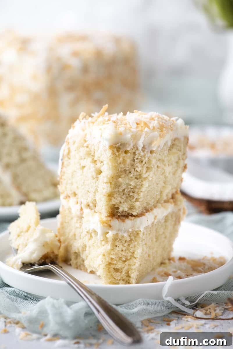Homemade Coconut Cake That Tastes Like Real Coconut 4 Two slices of coconut cake with white frosting are stacked on a white plate, topped with toasted coconut. A fork with a bite of cake rests beside the slices. The background includes more cake and a soft turquoise cloth.