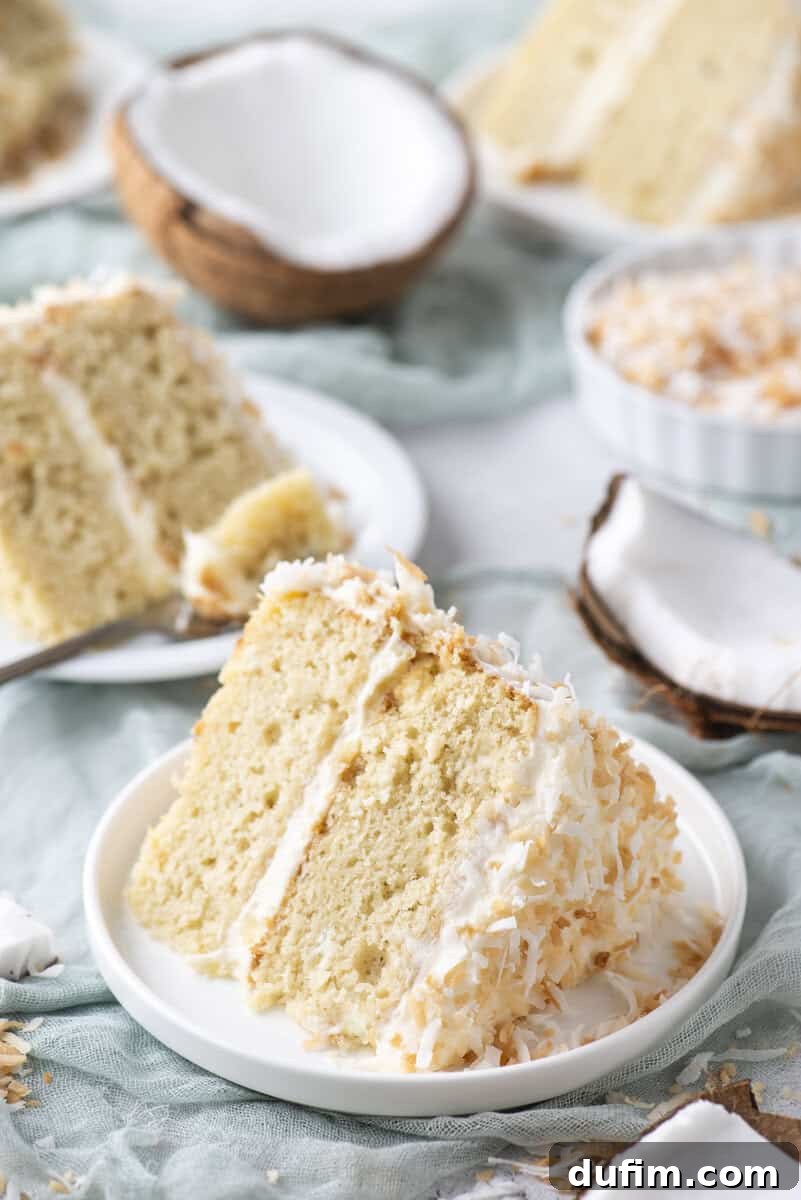 Homemade Coconut Cake That Tastes Like Real Coconut 6 A slice of coconut cake with white frosting and shredded coconut on a plate, surrounded by coconut halves and more cake slices in the background.