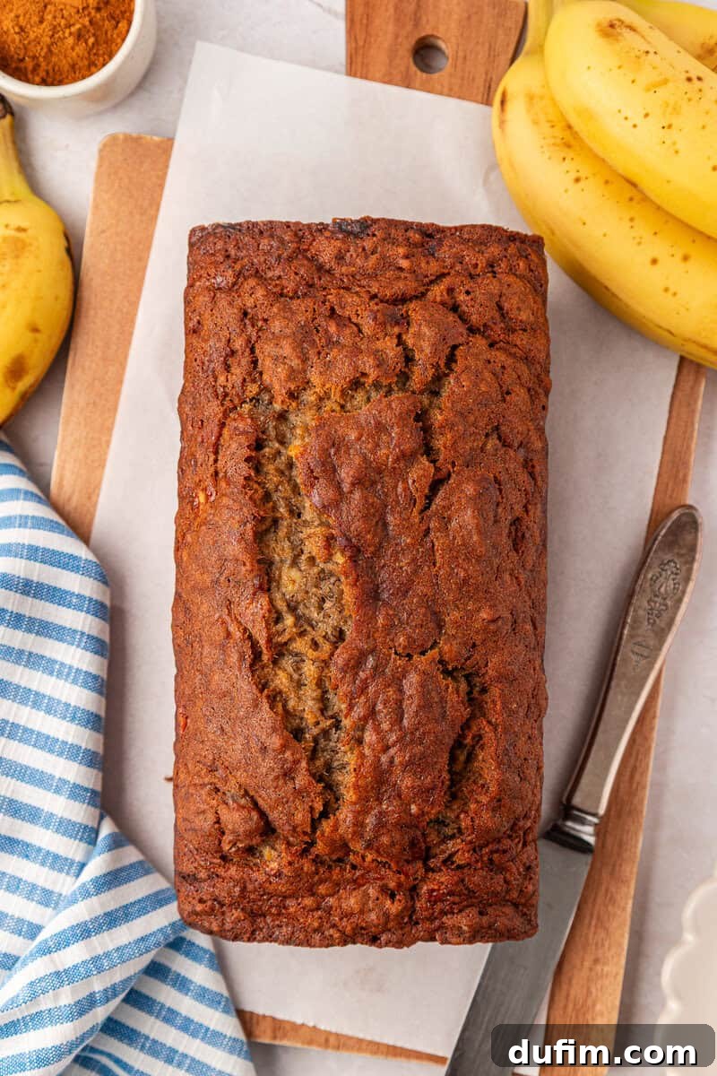 Moist Gluten-Free Banana Bread Recipe 2 A loaf of banana bread sits on parchment paper, surrounded by bananas, a knife, a wooden board, and a blue-striped cloth.