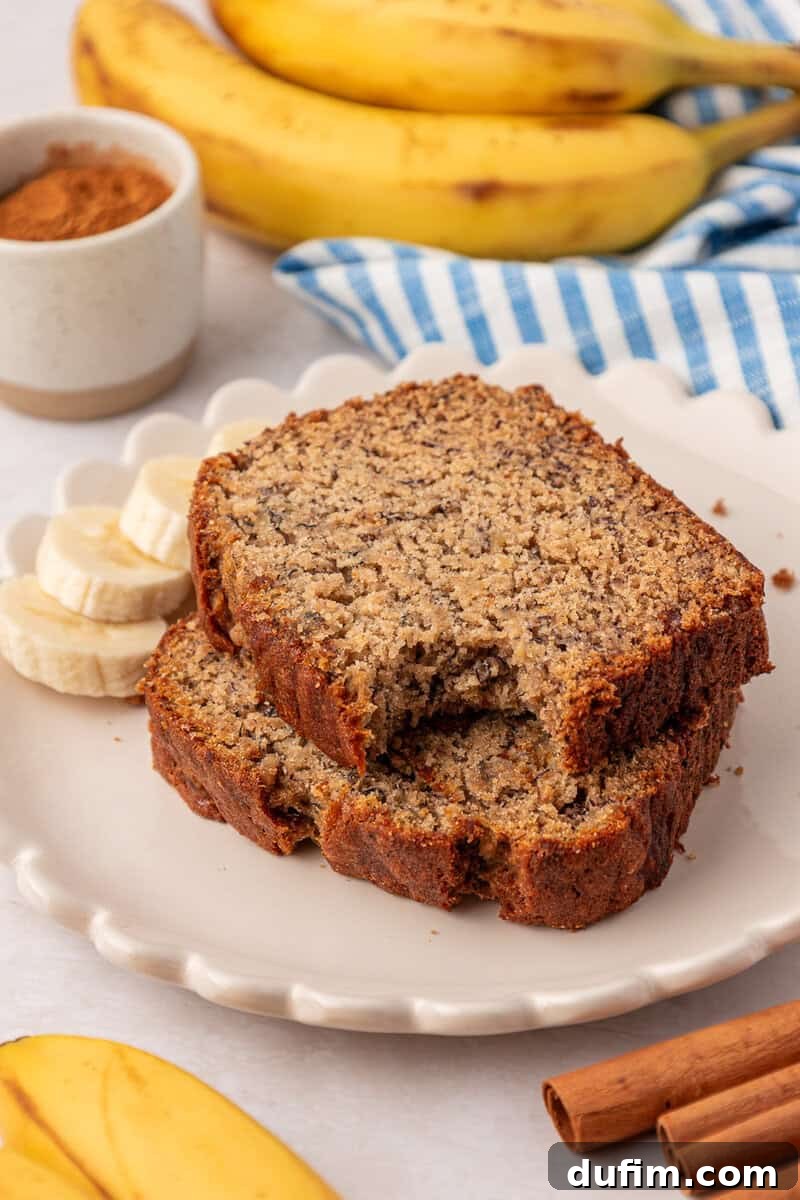 Moist Gluten-Free Banana Bread Recipe 14 Two slices of banana bread on a plate with banana slices, cinnamon sticks, whole bananas, and a bowl of cinnamon in the background.