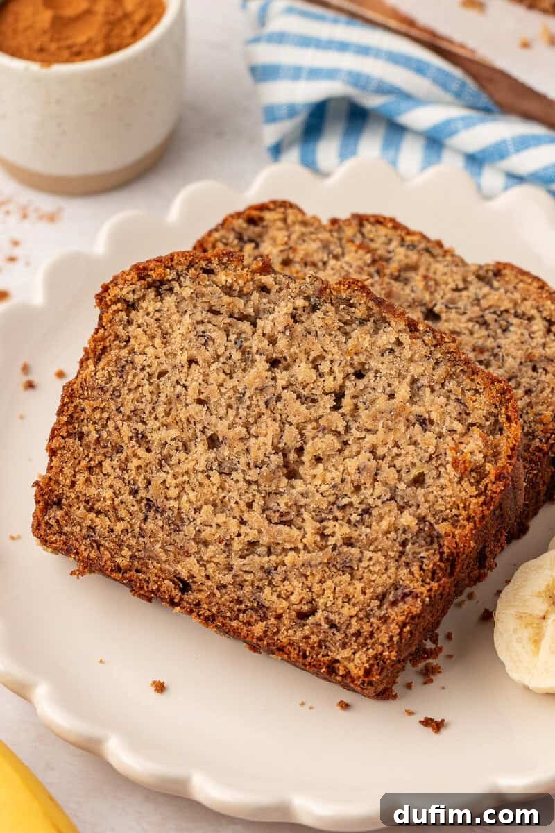 Moist Gluten-Free Banana Bread Recipe 15 Two slices of banana bread sit on a white scalloped plate, with a few banana slices nearby. A small bowl of cinnamon and a blue striped cloth are in the background.