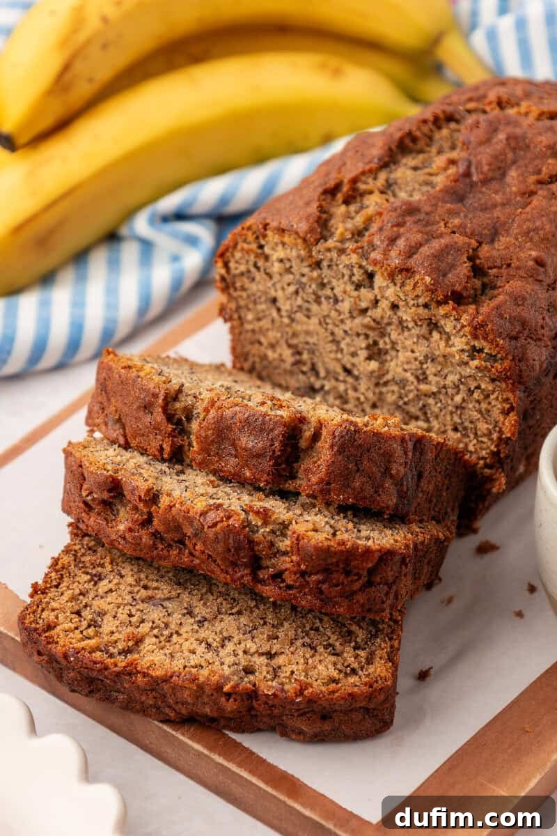 Moist Gluten-Free Banana Bread Recipe 16 A sliced loaf of banana bread sits on a wooden cutting board, with fresh bananas and a blue-striped cloth in the background.