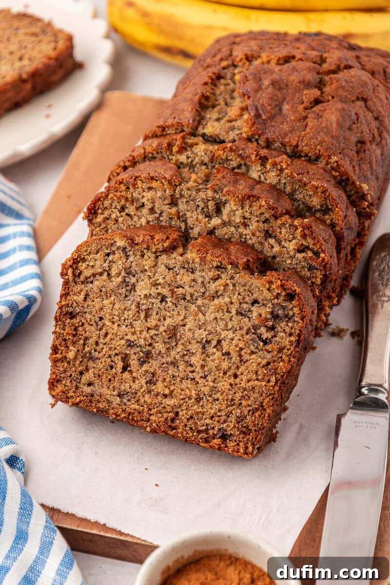 Moist Gluten-Free Banana Bread Recipe 17 A loaf of banana bread sliced on a cutting board, with a knife beside it and a bunch of bananas in the background.