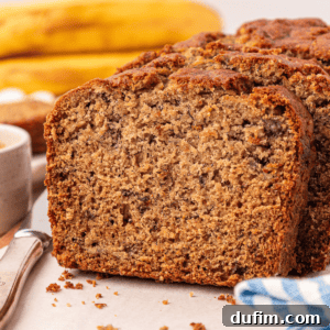 A close-up of sliced banana bread on a white surface, with bananas and a knife in the background. The bread looks moist, dense, and has a golden-brown crust.