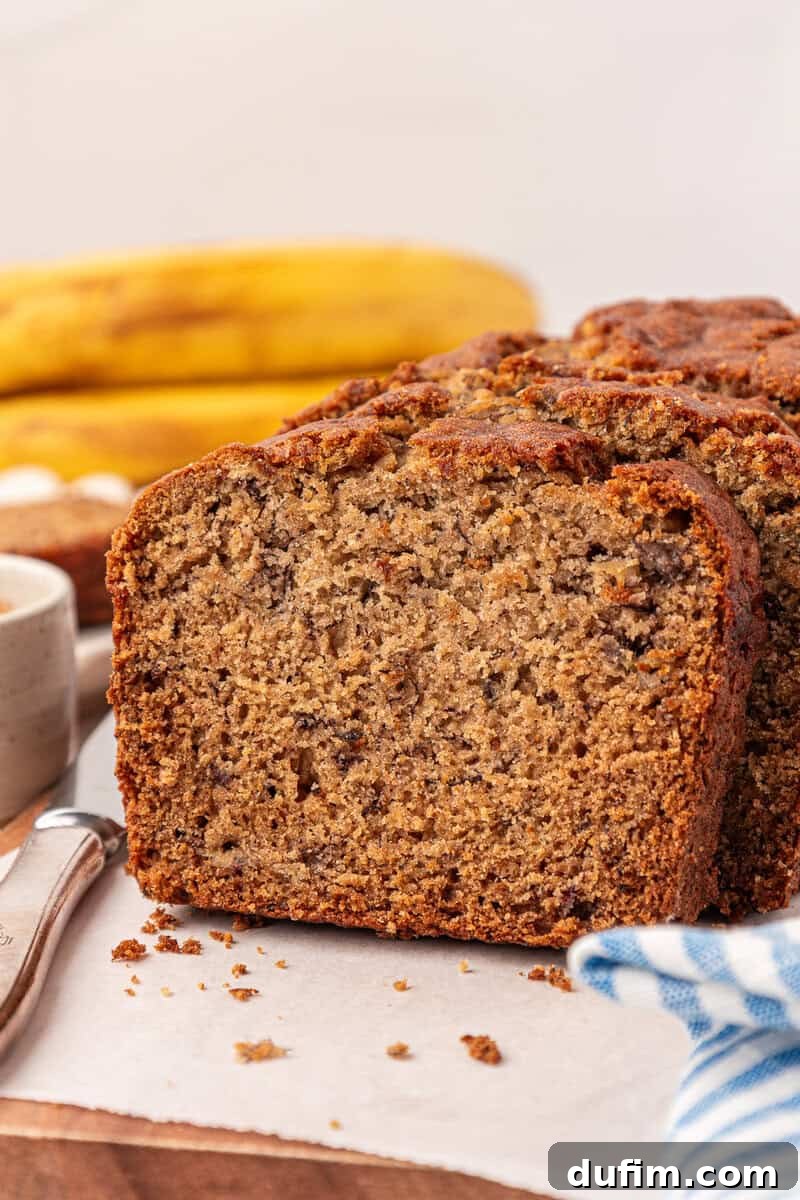 Moist Gluten-Free Banana Bread Recipe 5 A close-up of sliced banana bread on parchment paper, with ripe bananas and a cup in the background. A blue and white cloth is partially visible in the foreground.