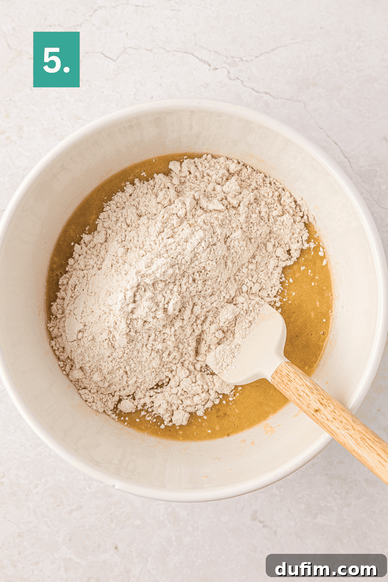 Moist Gluten-Free Banana Bread Recipe 10 A mixing bowl with flour sitting on top of a wet batter, being stirred with a white spatula.