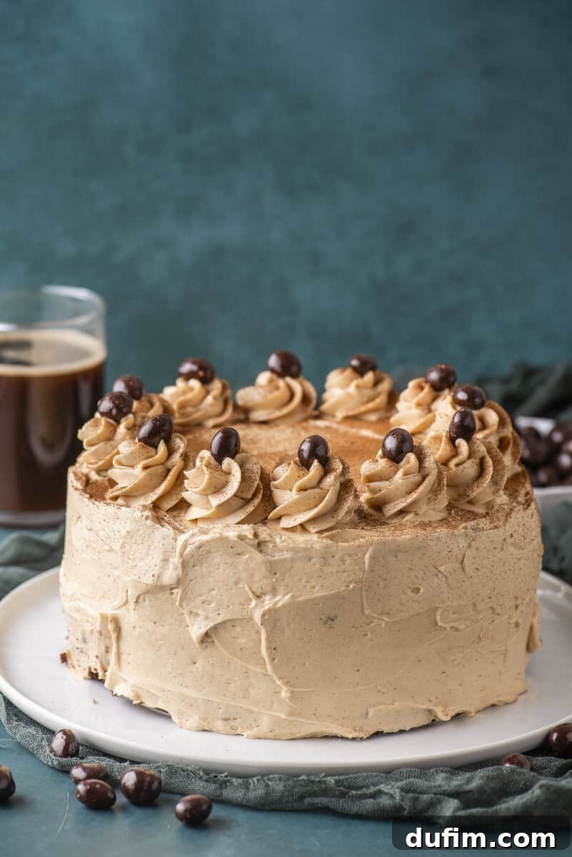 Decadent Mocha Cake Recipe for Coffee Lovers 2 Round cake with light brown frosting, decorated with swirls of frosting and chocolate-covered coffee beans on top, sits on a white plate. There is a glass of coffee and scattered beans nearby, set against a blue background.