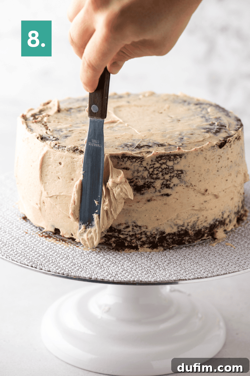 Decadent Mocha Cake Recipe for Coffee Lovers 14 A hand uses a spatula to spread brown frosting on a round cake placed on a white cake stand. A green box with the number 8 appears in the top left corner.