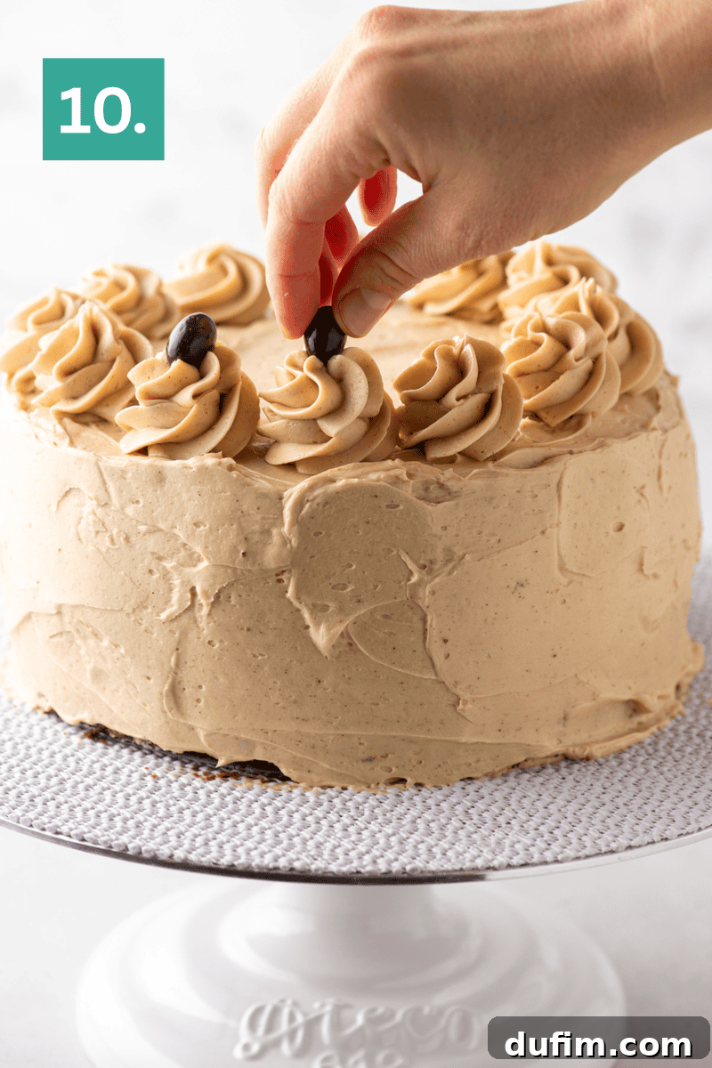 Decadent Mocha Cake Recipe for Coffee Lovers 16 A hand places a coffee bean on top of a swirl of frosting on a round cake with light brown icing, displayed on a white cake stand. A green box with the number 10 appears in the upper left corner.
