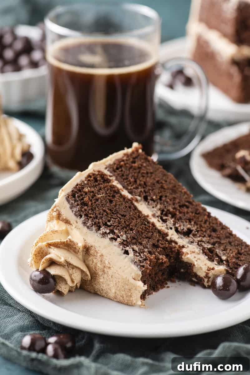 Decadent Mocha Cake Recipe for Coffee Lovers 19 A slice of chocolate layer cake with mocha frosting sits on a white plate, with chocolate-covered coffee beans beside it. A clear mug of black coffee and more cake slices are in the background.
