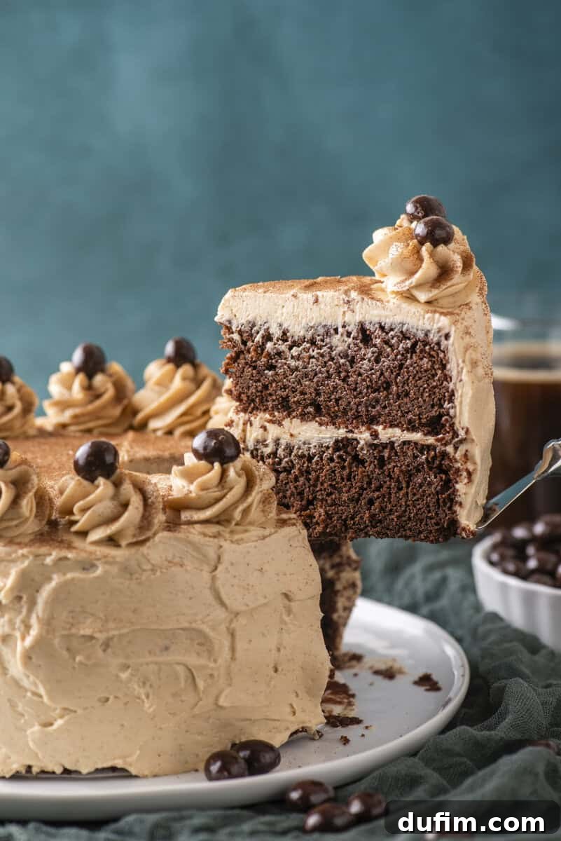 Decadent Mocha Cake Recipe for Coffee Lovers 3 A slice of chocolate layer cake with light brown frosting is being lifted from a decorated cake, topped with swirls of frosting and dark round candies. A cup and a bowl of similar candies are in the background.