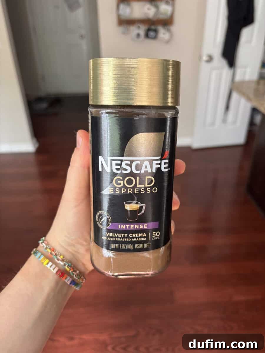 Decadent Mocha Cake Recipe for Coffee Lovers 5 A hand holds a jar of Nescafé Gold Espresso Intense coffee. The jar has a gold lid and a black label with a coffee cup image. The background shows a kitchen with wooden floors and white doors.
