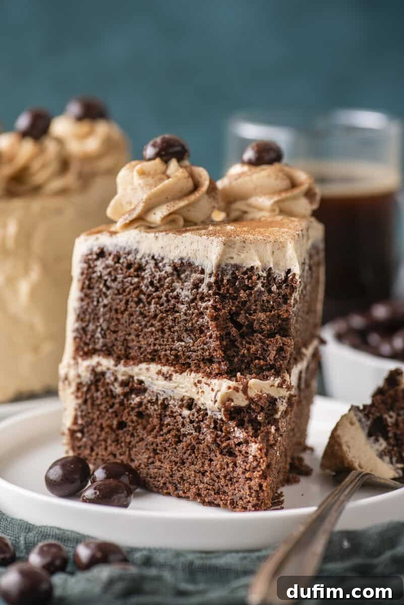 Decadent Mocha Cake Recipe for Coffee Lovers 6 A slice of chocolate cake with creamy frosting, topped with swirls and chocolate-coated coffee beans, on a white plate with a fork. Blurred coffee and more cake are in the background.