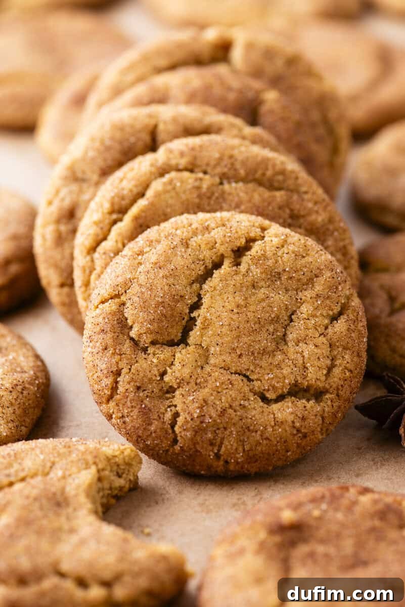 A close-up of several golden-brown, sugar-coated cookies stacked and scattered on a light surface. One cookie in front displays a cracked top, with another partially eaten cookie nearby.