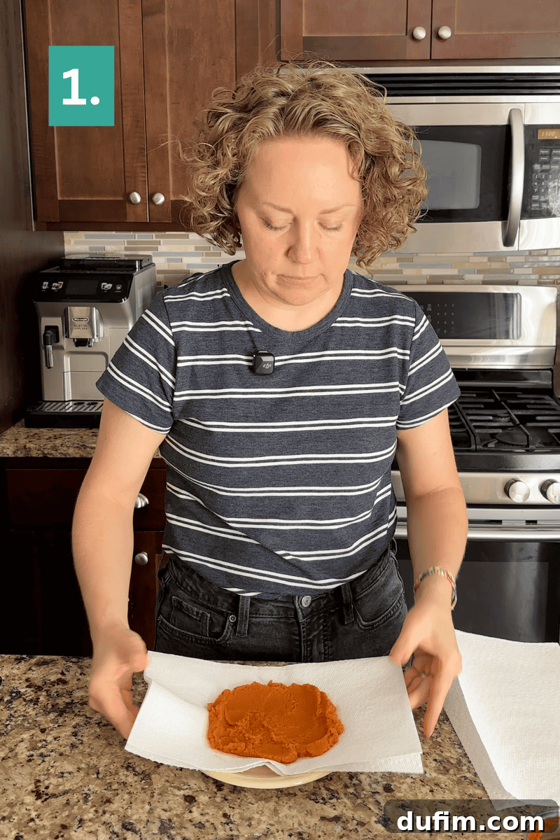 A person in a striped shirt presses pumpkin puree on a plate with paper towels in a kitchen. A green square with the number 1. appears in the top left corner.