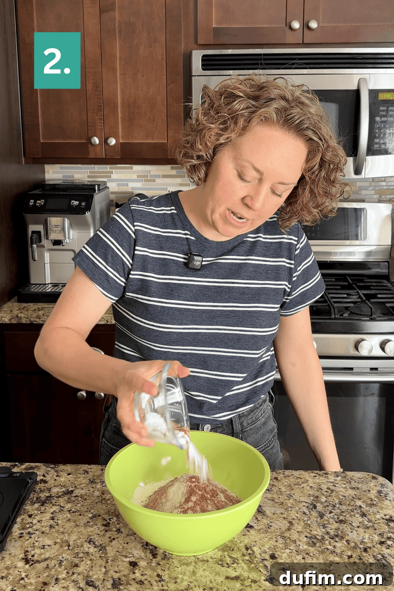 A person in a striped shirt pours a cup of white powder into a green mixing bowl with dry ingredients on a kitchen counter. A green box with 2. is in the top left corner.