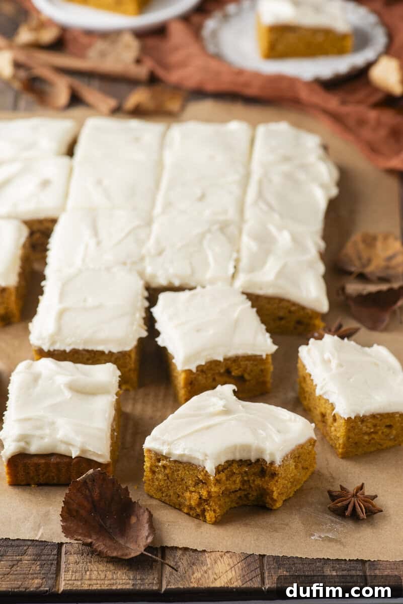 Pumpkin bars with white frosting cut into squares on parchment paper, surrounded by autumn leaves and spices. One bar in the foreground has a bite taken out of it.