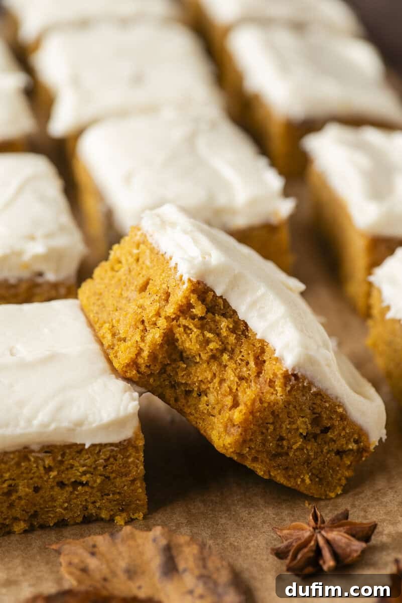 Pumpkin bars topped with creamy white frosting, arranged on brown parchment paper. One bar is propped up to show its moist texture. A star anise and a dry leaf sit nearby.